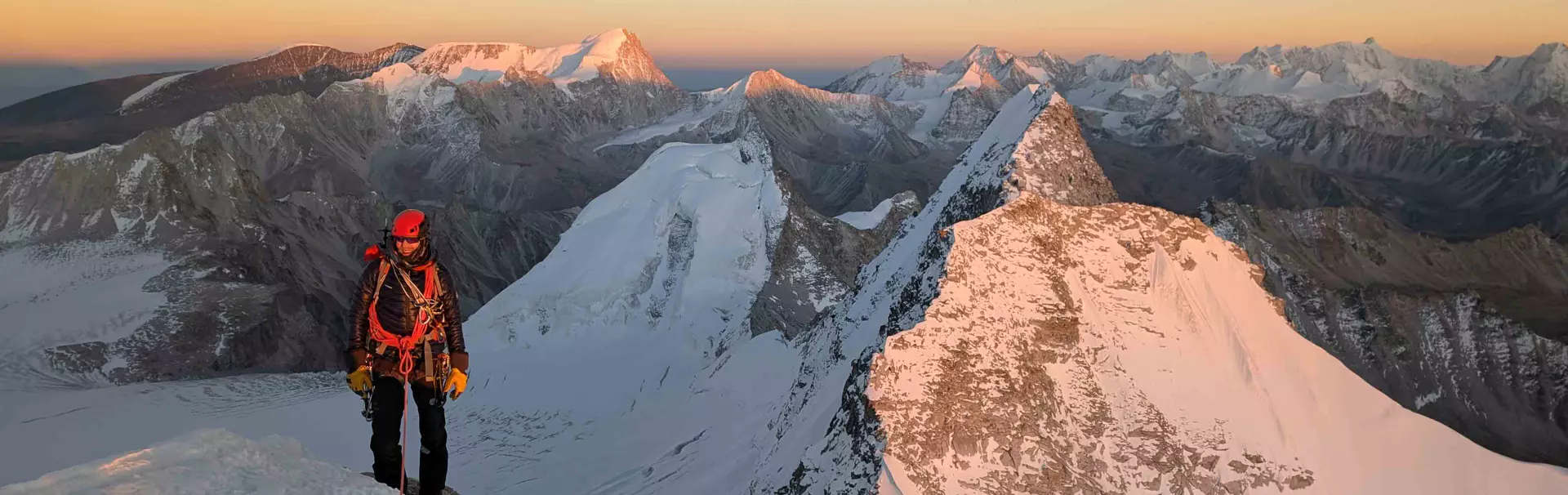 Een bergbeklimmer in winteruitrusting, met een klimgordel en een helm, staat bij zonsopgang op een besneeuwde bergkam, omringd door ruige, met sneeuw bedekte bergtoppen en indrukwekkende landschappen.